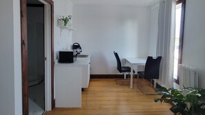 Cocina compacta con cubierta blanca, un lavamanos y un microondas. Hay un área de comedor con una mesa y dos sillas. El piso es de madera y las paredes son blancas.