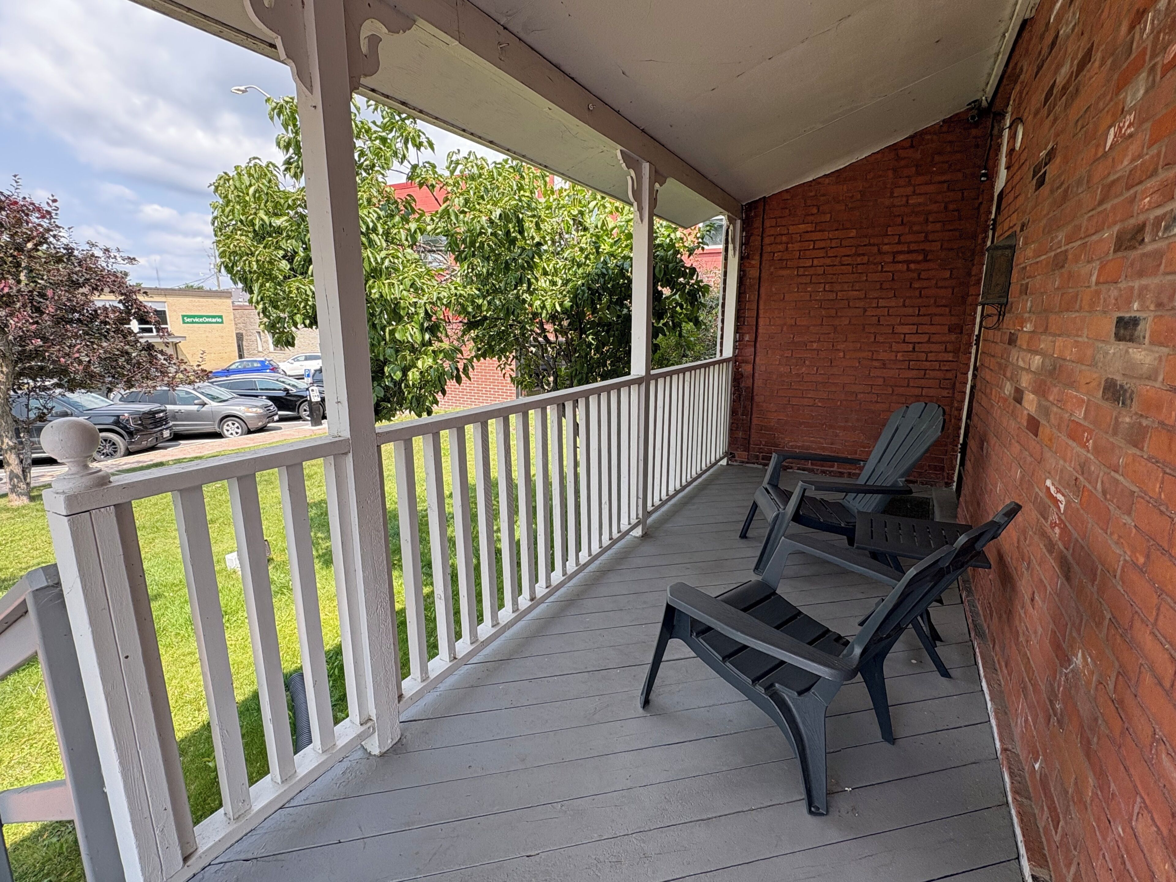 A covered porch with a white railing, two Adirondack chairs, and a brick wall.