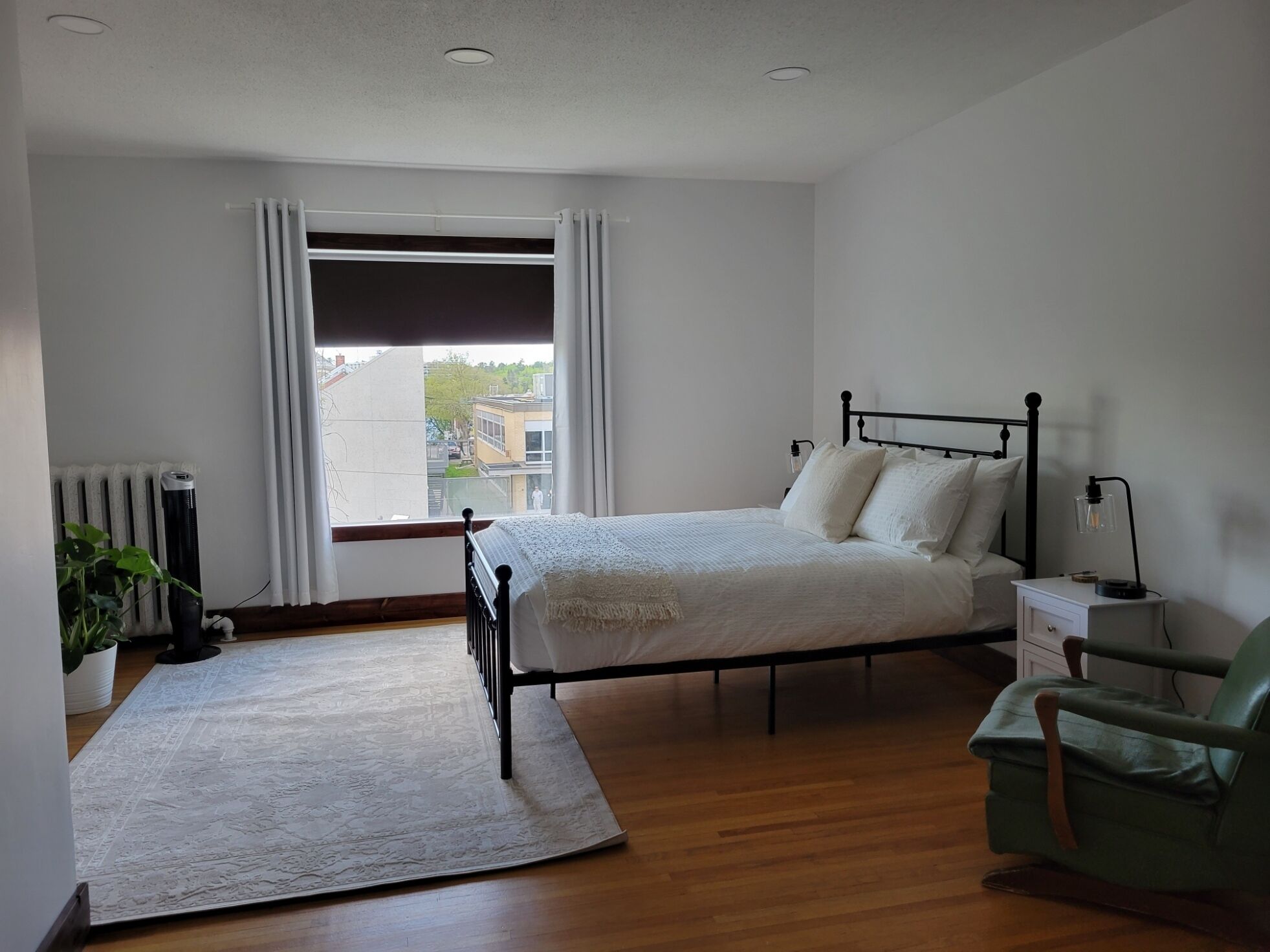 A bedroom with a bed, a nightstand, a chair, a radiator, and a window with curtains.