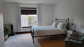 A bedroom with a bed, a nightstand, a chair, a radiator, and a window with curtains.