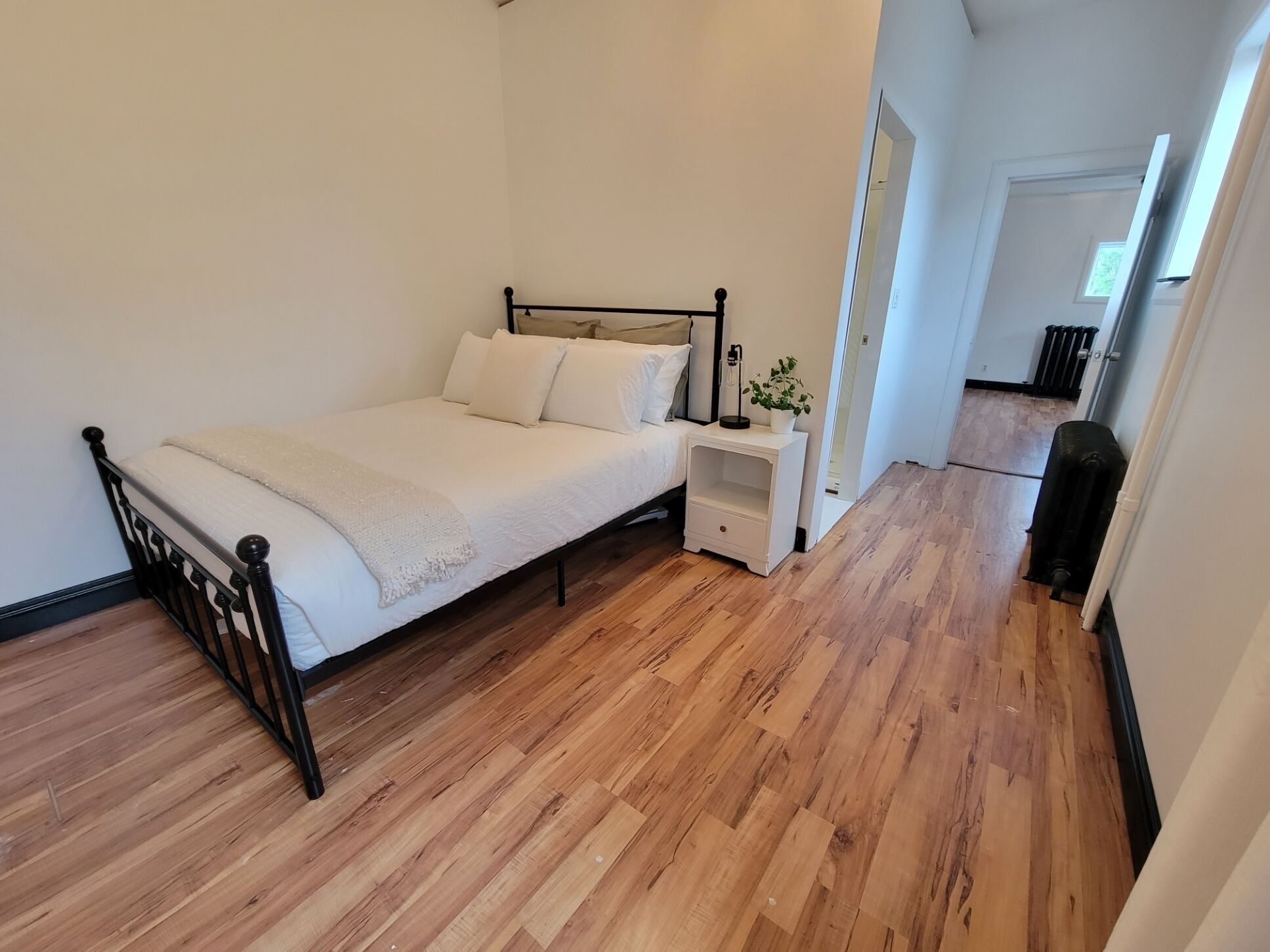 A bedroom with a wooden floor, a bed with white bedding, a nightstand with a plant, and a radiator.