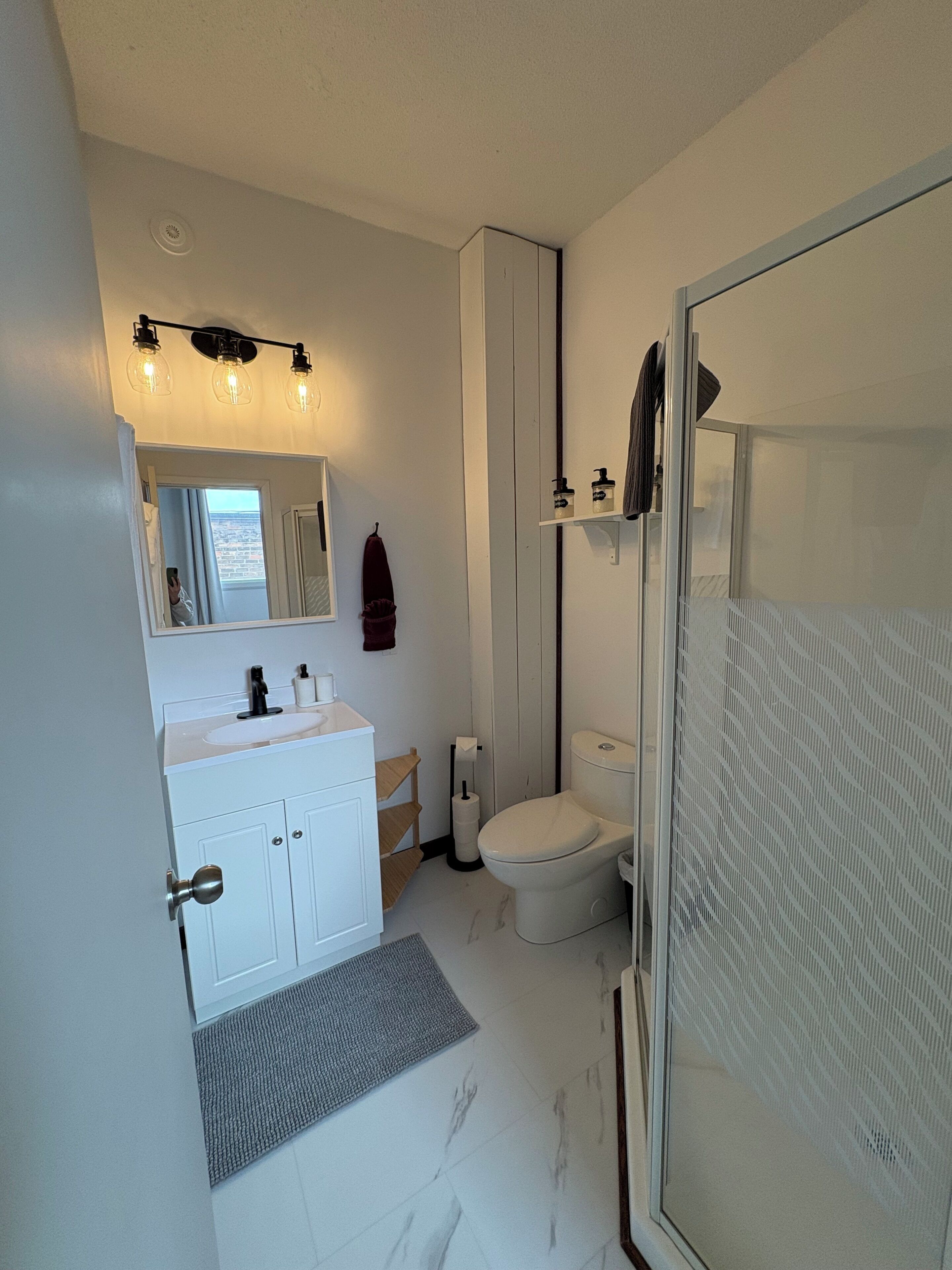 Un bagno moderno con lavandino, specchio e water bianchi. È presente una cabina doccia in vetro e un piccolo ripiano con articoli da toilette.