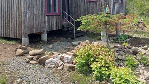 A rustic wooden cabin with two windows, a small porch, and a garden area with rocks and plants. - Fundy Fantasy Oceanfront Cabin. Offgrid romantic getaway. (Granville Ferry)