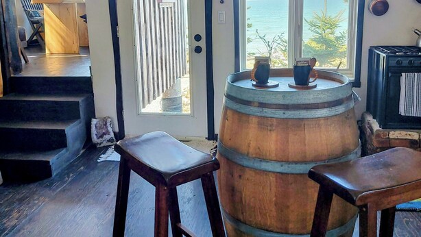 A rustic interior with a wooden barrel, two stools, a staircase, and a view of the sea through a large window. - Fundy Fantasy Oceanfront Cabin. Offgrid romantic getaway. (Granville Ferry)
