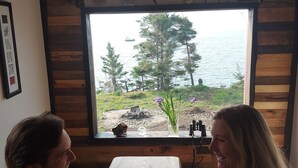 A wooden cabin with a large window overlooking a lake, a dining table with plates of food, and binoculars on the windowsill. - Fundy Fantasy Oceanfront Cabin. Offgrid romantic getaway. (Granville Ferry)
