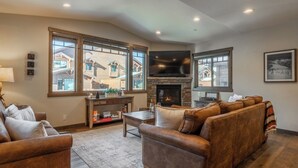 A cozy living room with a fireplace, leather sofas, a coffee table, and large windows offering a view of the outside.