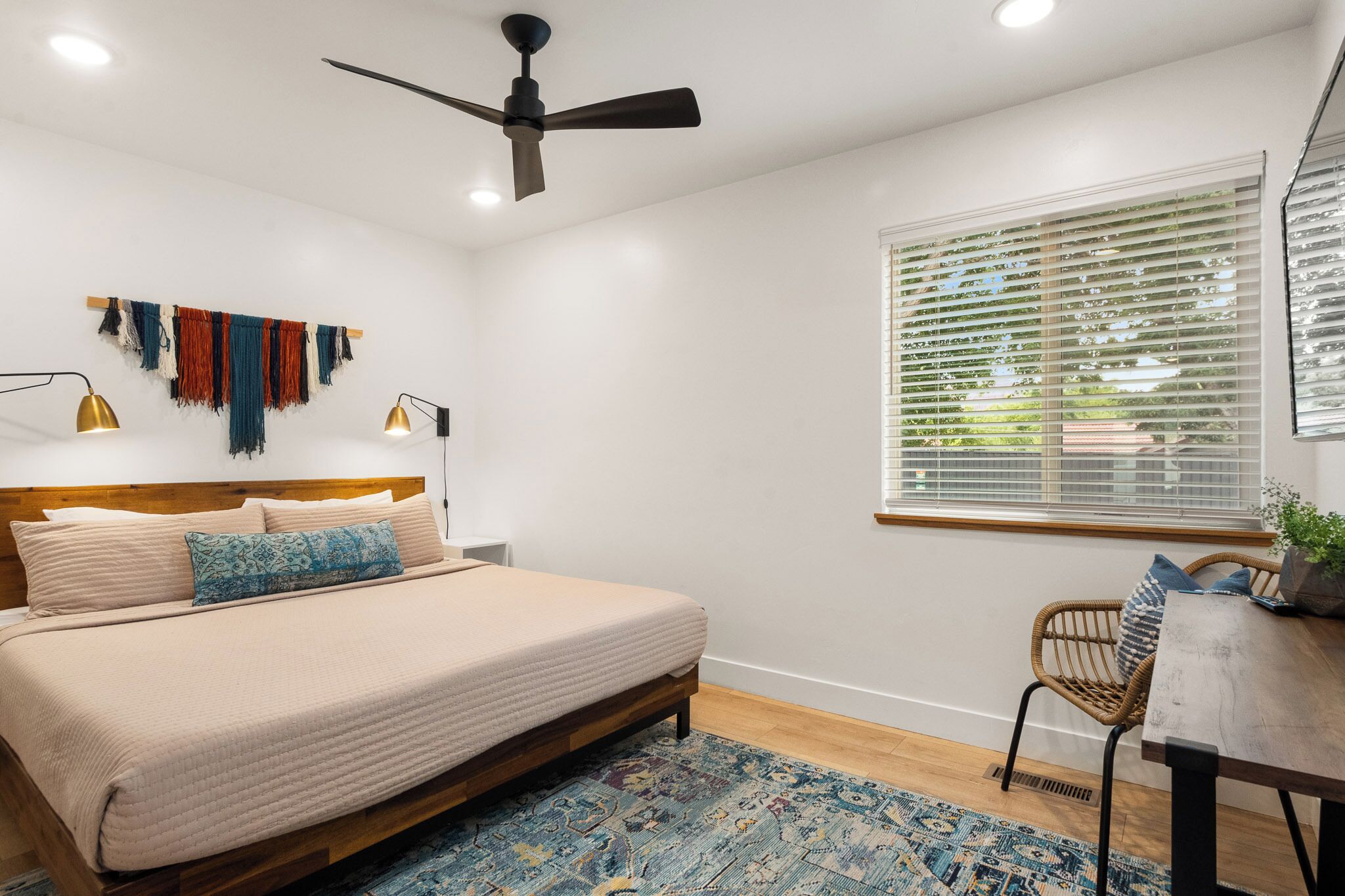 A bedroom with a bed, a ceiling fan, a window with blinds, a bedside table with a lamp, and a chair.