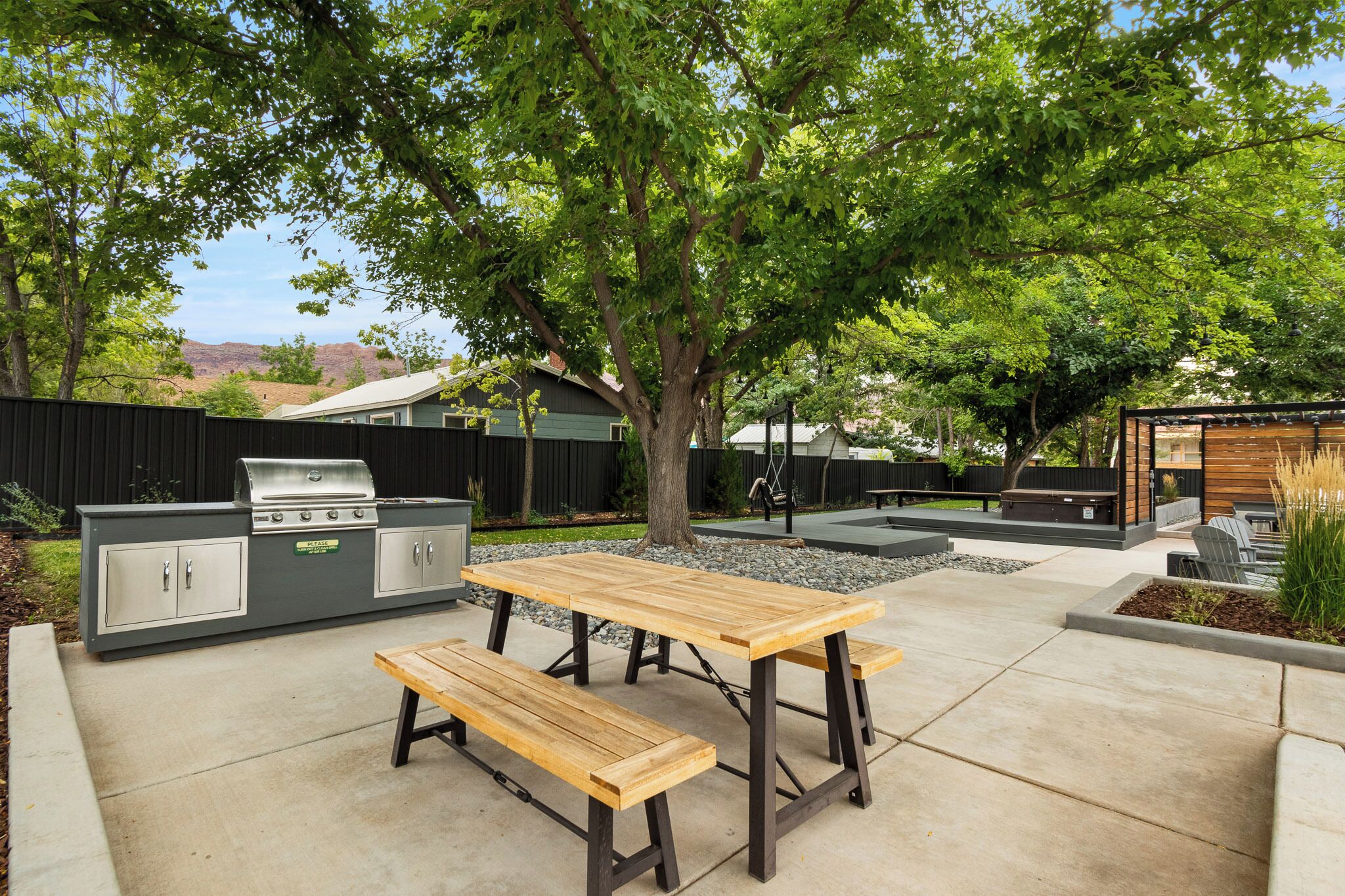 An outdoor patio with a wooden table and benches, a barbecue grill, and a fenced area with a tree.