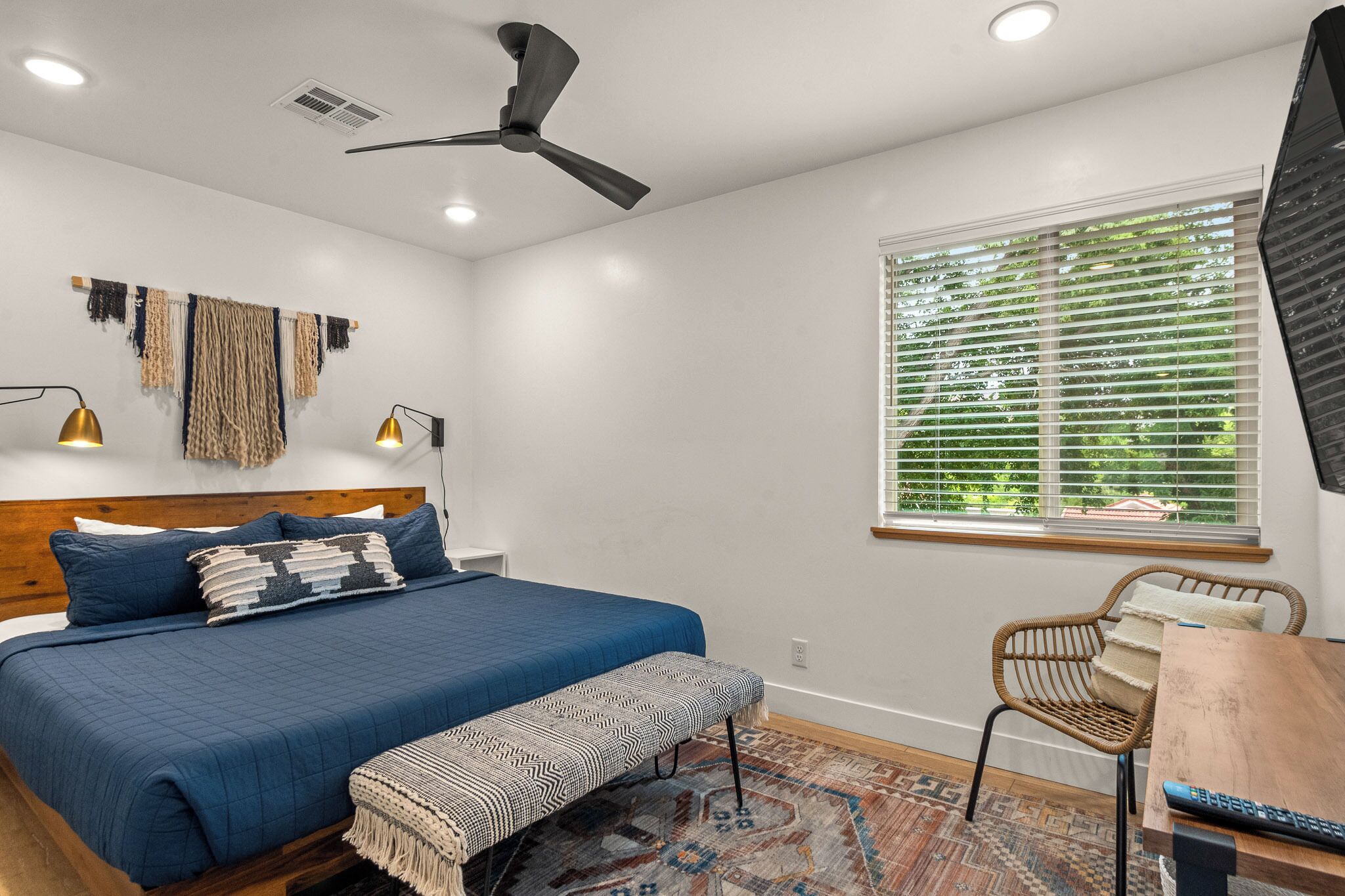 A bedroom with a bed, a desk, a chair, a ceiling fan, and a window with blinds.
