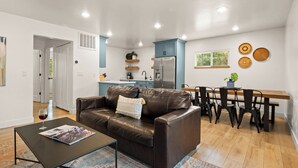 Ein modernes Wohnzimmer mit einer Ledersofa, einem Esstisch mit Stühlen, einer Küchenzeile mit blauen Schränken und einem kleinen Tisch mit einer Pflanze.