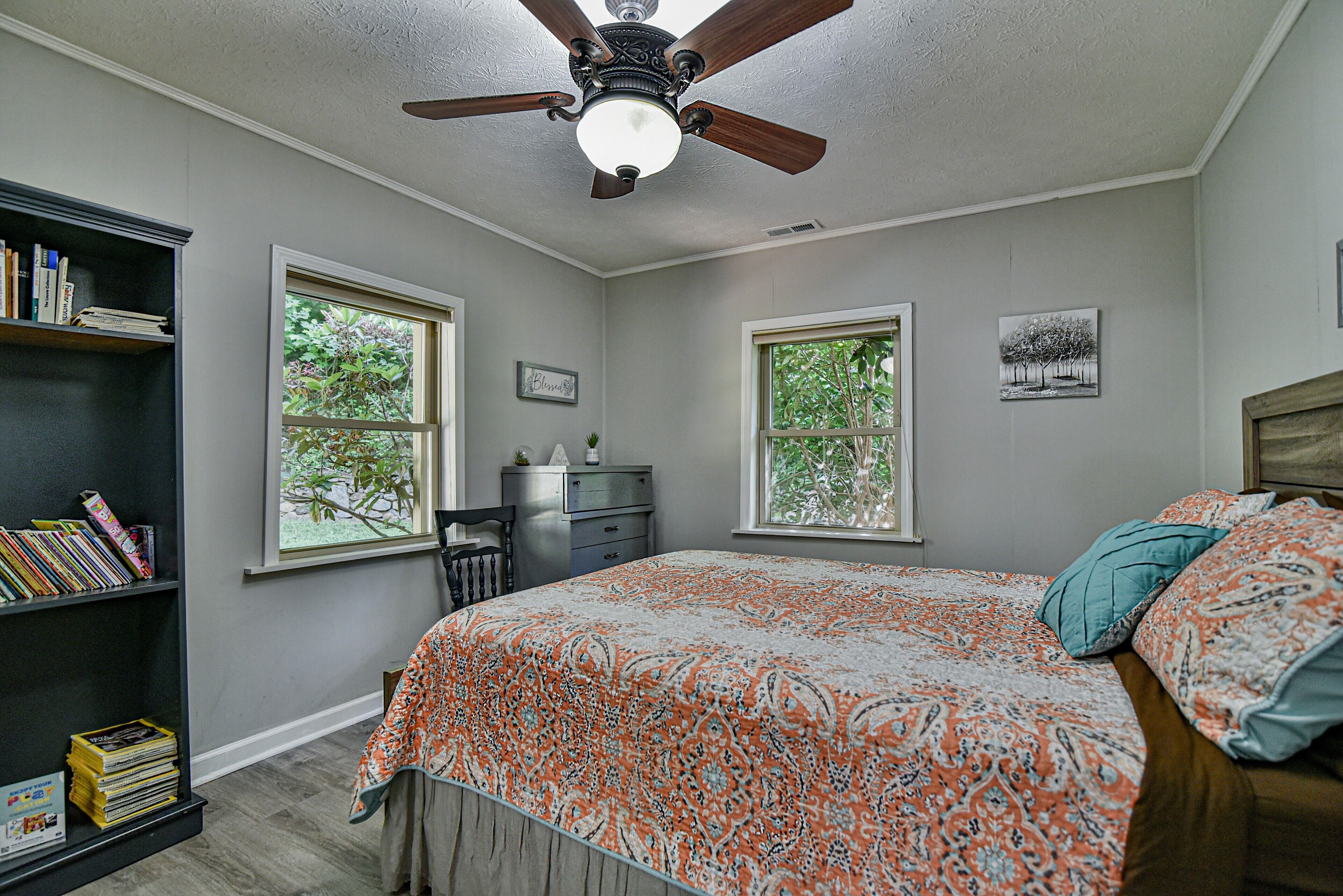 Un dormitorio con cama, estantería, escritorio y dos ventanas.