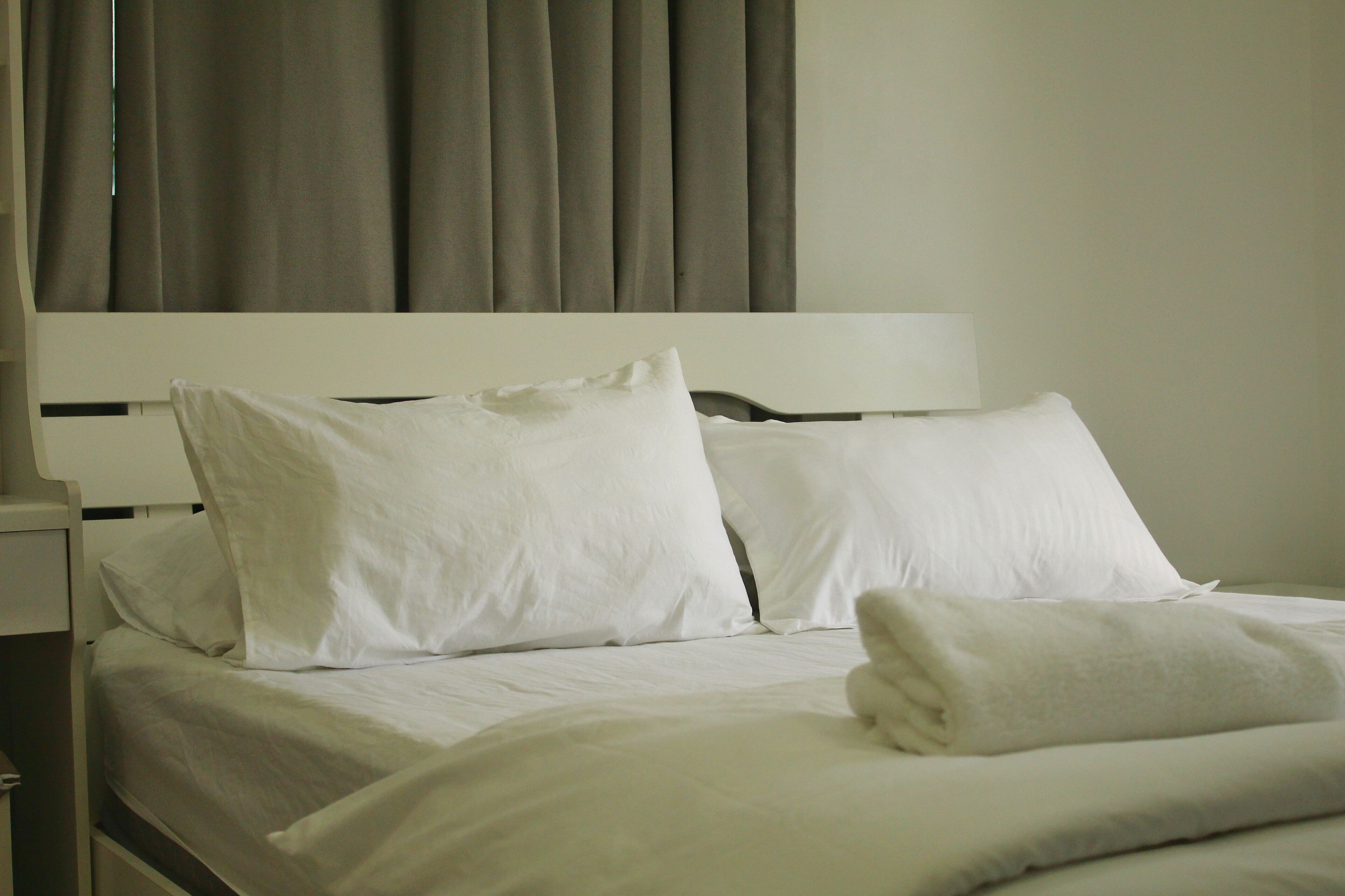 A neatly made bed with white linens and pillows, a white headboard, and a window with curtains.