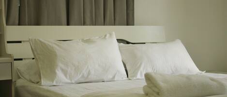 A neatly made bed with white linens and pillows, a white headboard, and a window with curtains.