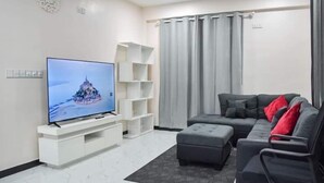 Ein modernes Wohnzimmer mit Flachbildfernseher, einem grauen Ecksofa mit roten Kissen, einer weißen Unterhaltungsanlage und grauen Vorhängen.
