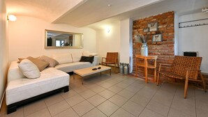 Een woonkamer met een witte bank, een salontafel en twee rieten stoelen. Er is een bakstenen muur met een plank en een klein tafeltje met een vaas.