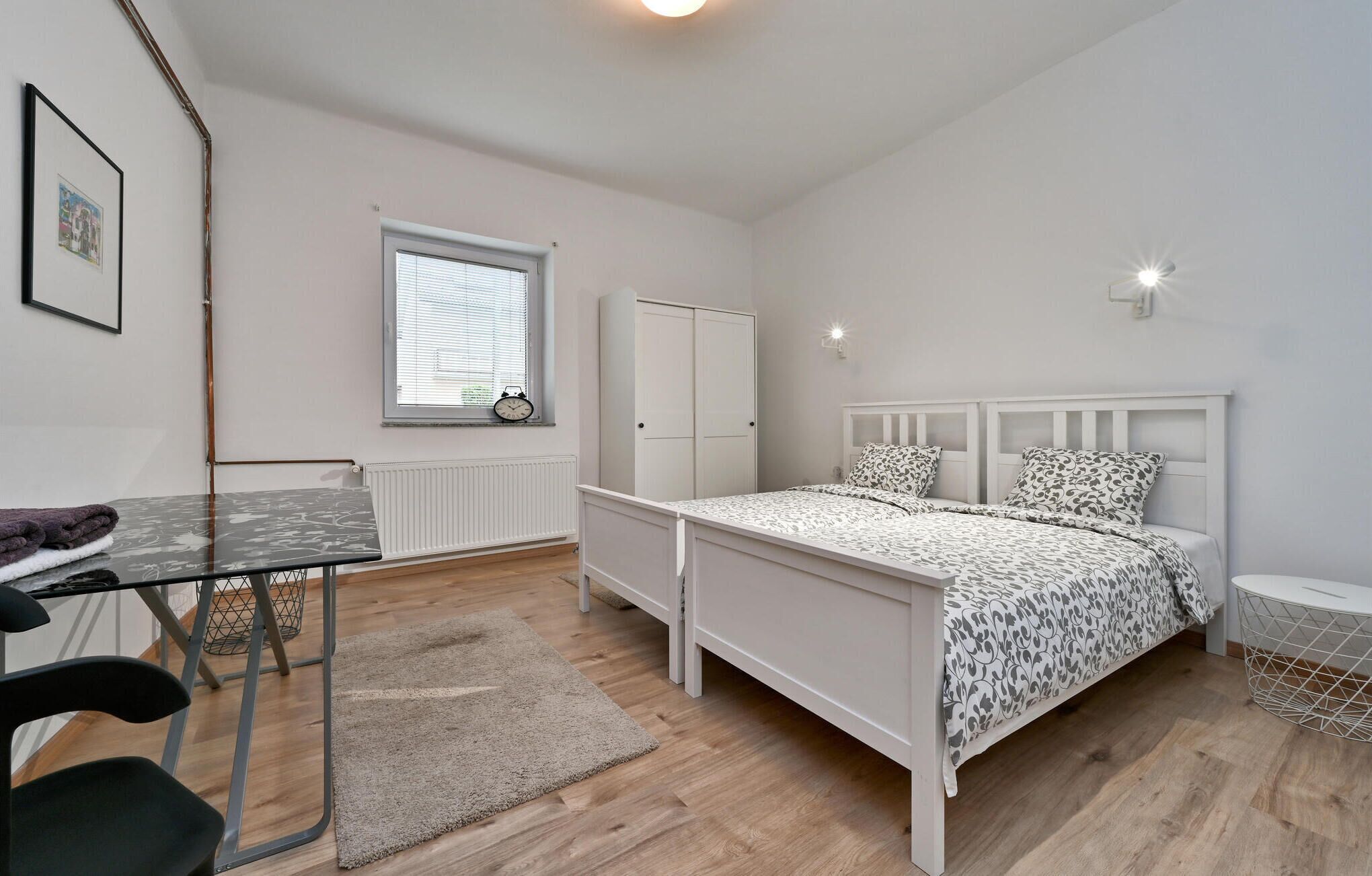 A bedroom with a bed, a wardrobe, a radiator, a window with blinds, a desk with a chair, and a small table with a chair.
