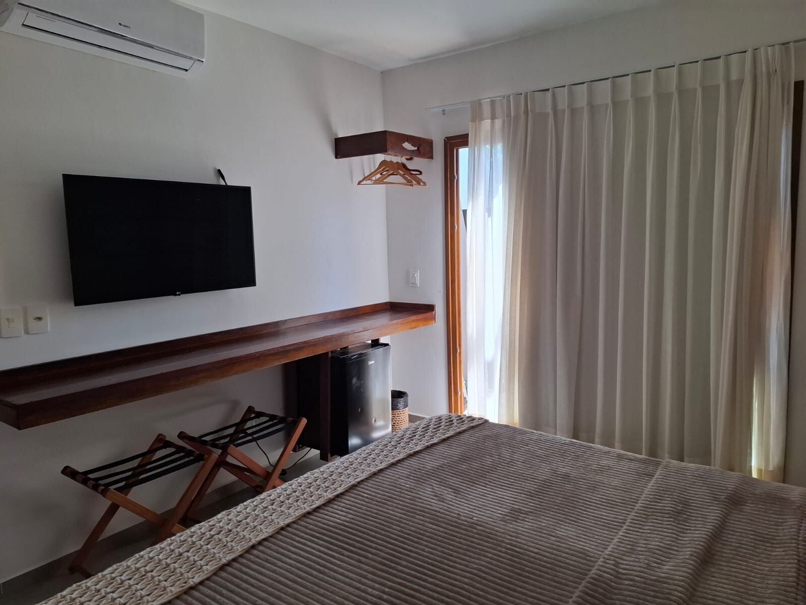 Una habitación de hotel moderna con un televisor de pantalla plana montado en la pared, un banco de madera, un radiador y un gran ventanal con cortinas.