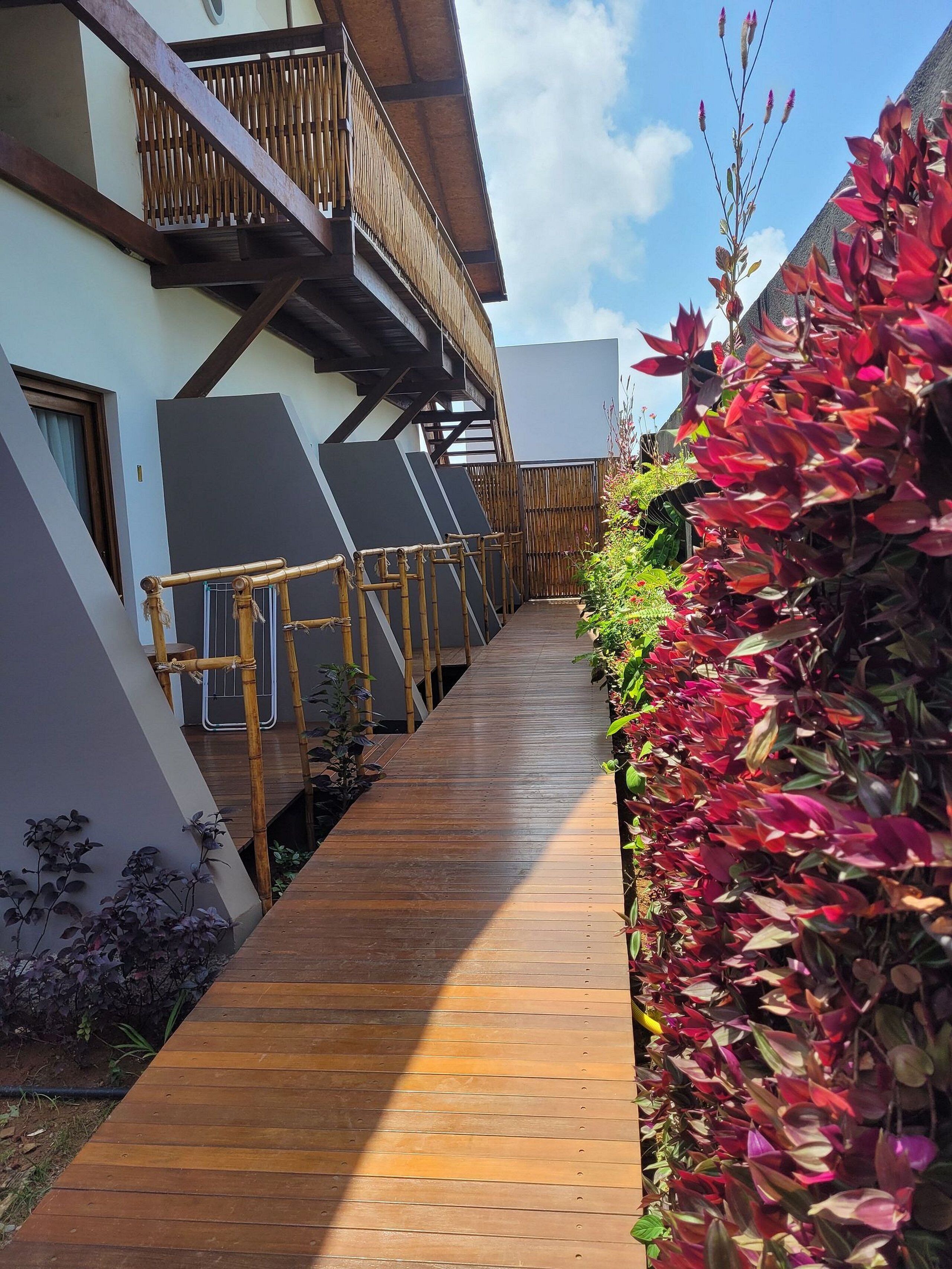 A wooden pathway leading to a building with a balcony, surrounded by lush greenery and a clear sky.