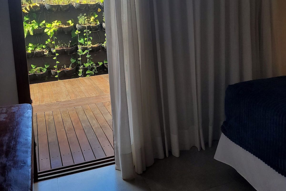 Une chambre avec un lit, un plancher en bois et une vue sur un jardin, visible à travers une fenêtre ornée de rideaux blancs.