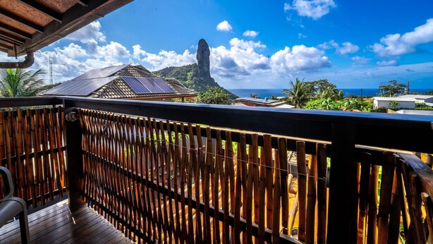 Eine Holzterrasse mit Bambuszaun, Solaranlage auf einem Gebäude und freier Blick auf Berg und Meer.
