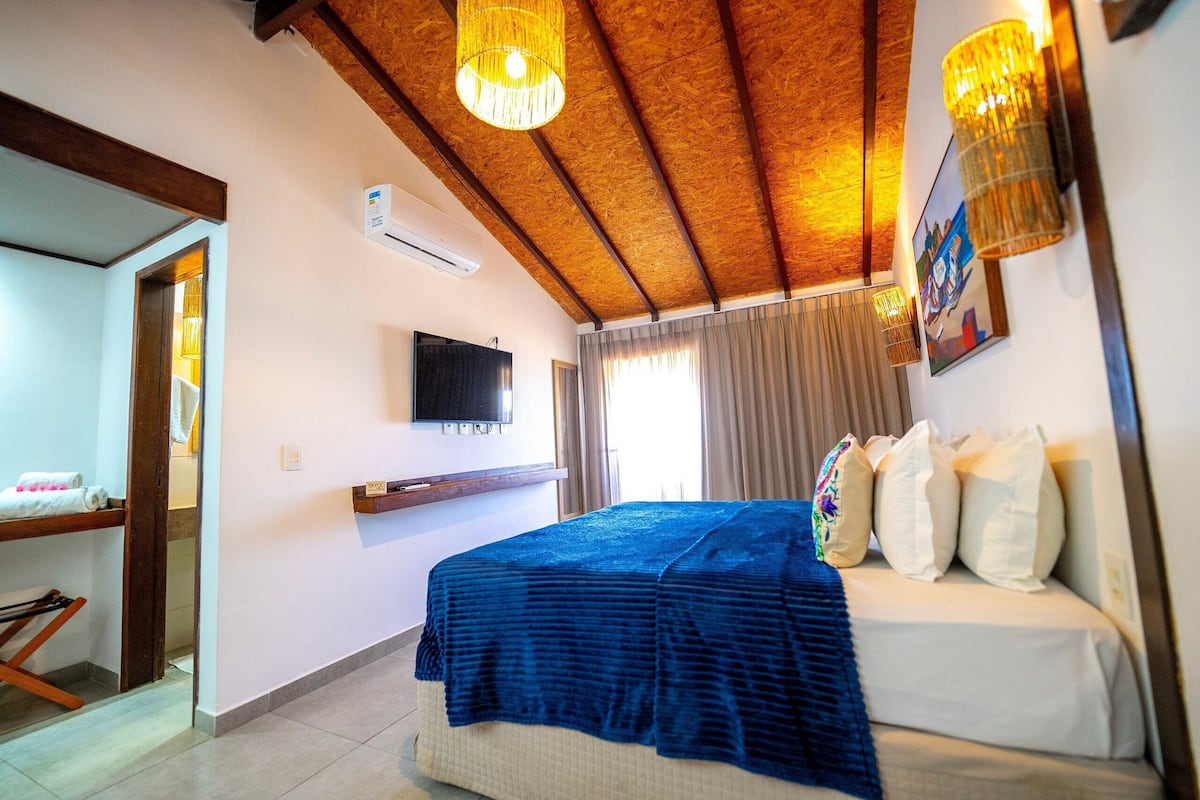 Une chambre à coucher comprenant un lit, un téléviseur fixé au mur, un ventilateur de plafond et une fenêtre avec des rideaux.