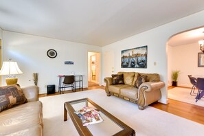 A living room with a beige sofa, a coffee table with magazines, a painting on the wall, and a dining area in the background.