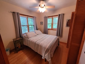 Une chambre à coucher avec un lit, deux fenêtres équipées de stores, un ventilateur de plafond, une table de chevet et un plancher en bois.