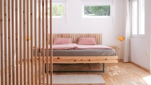 Een minimalistische slaapkamer met een houten bed, twee ramen en uitzicht op groen.