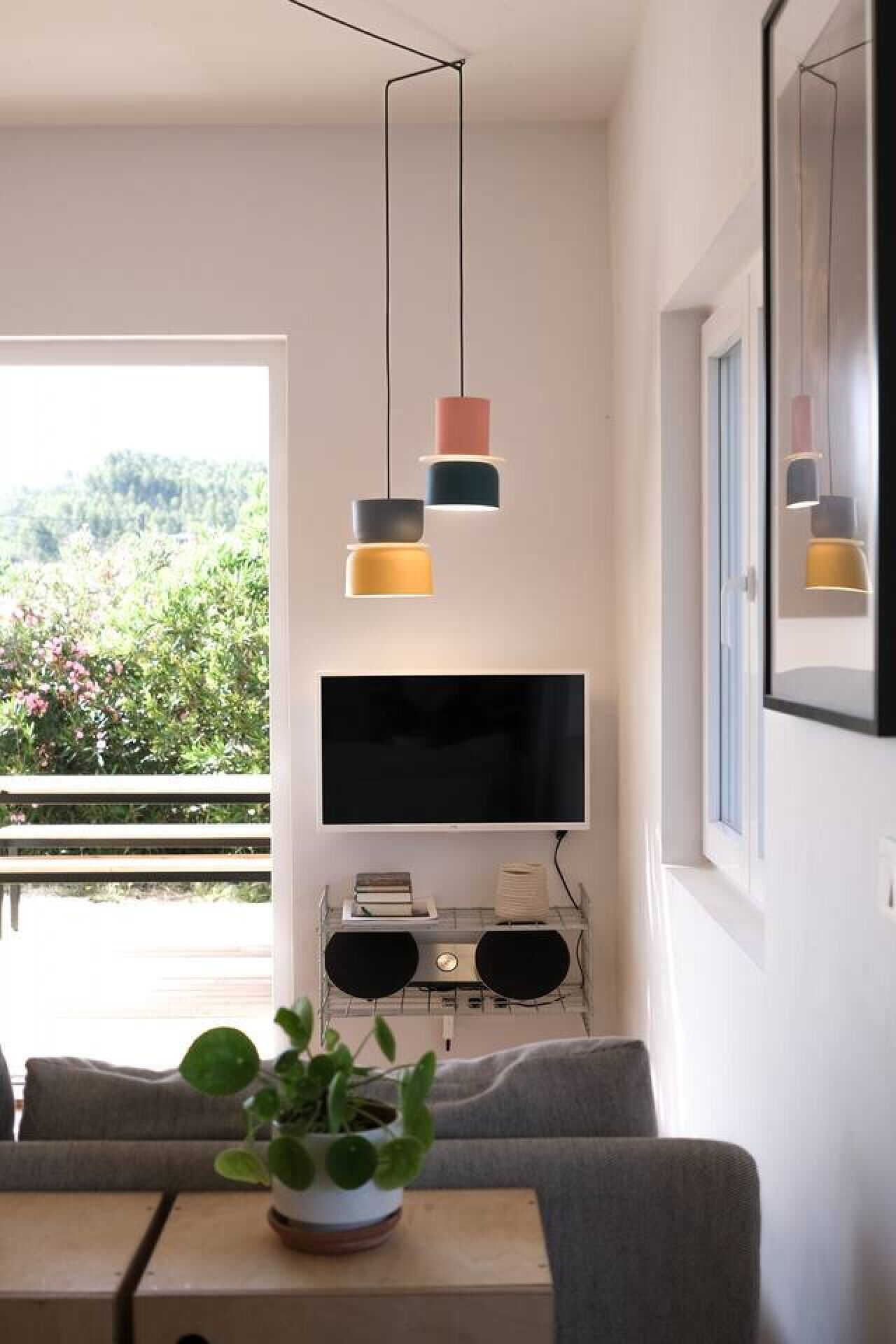 A modern living room with a flat-screen TV, a sofa, a dining table with a plant, and a balcony with a view of greenery.