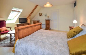 Ein Schlafzimmer mit schrägen Wänden, einem Bett mit blattbedeckter Bettdecke, einem Holznachttisch, einem roten Sessel, einem kleinen Tisch mit einem Fernseher und einem Fenster mit Blick auf Grünflächen.