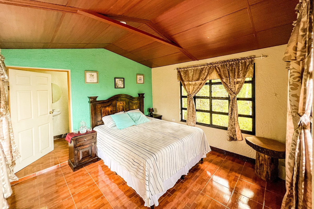A bedroom with a wooden ceiling, a bed with striped bedding, a nightstand, a window with curtains, and a door.