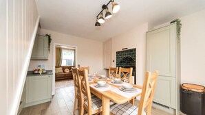 A dining area with a wooden table set for four, surrounded by wooden chairs with plaid cushions. A kitchen area is visible to the left, with a green tiled backsplash and a white cabinet.