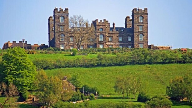 A large, historic building with multiple towers and a central courtyard, surrounded by lush green fields.