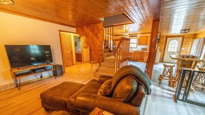 A cozy living room with a large flat-screen TV, leather sofa, and a staircase leading to a kitchen. - Secluded 3BR Cabin • Fire Pit • RV Parking • Near Tippy Dam & Big-M Trails (Wellston)