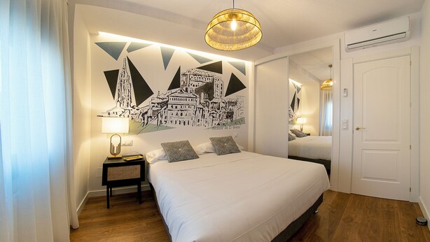 A hotel room with a large bed, a nightstand with a lamp, a wall mural, and a door. - ALCAZAR CERVANTES 5 COLLECTION (Toledo)