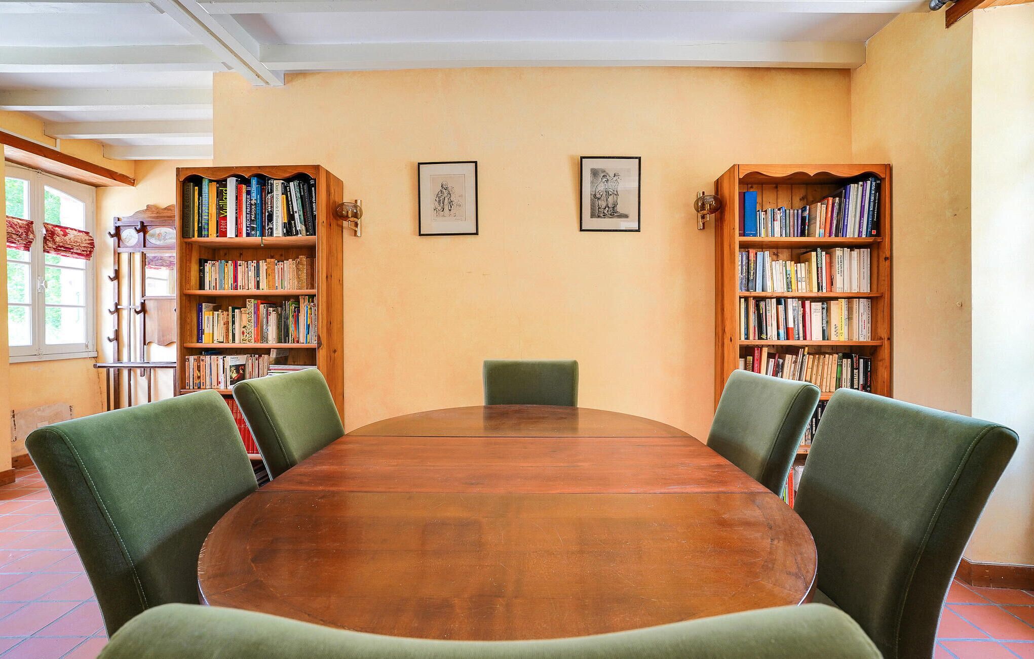 A library room with a round wooden table, green upholstered chairs, and bookshelves filled with books.