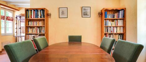 A library room with a round wooden table, green upholstered chairs, and bookshelves filled with books.