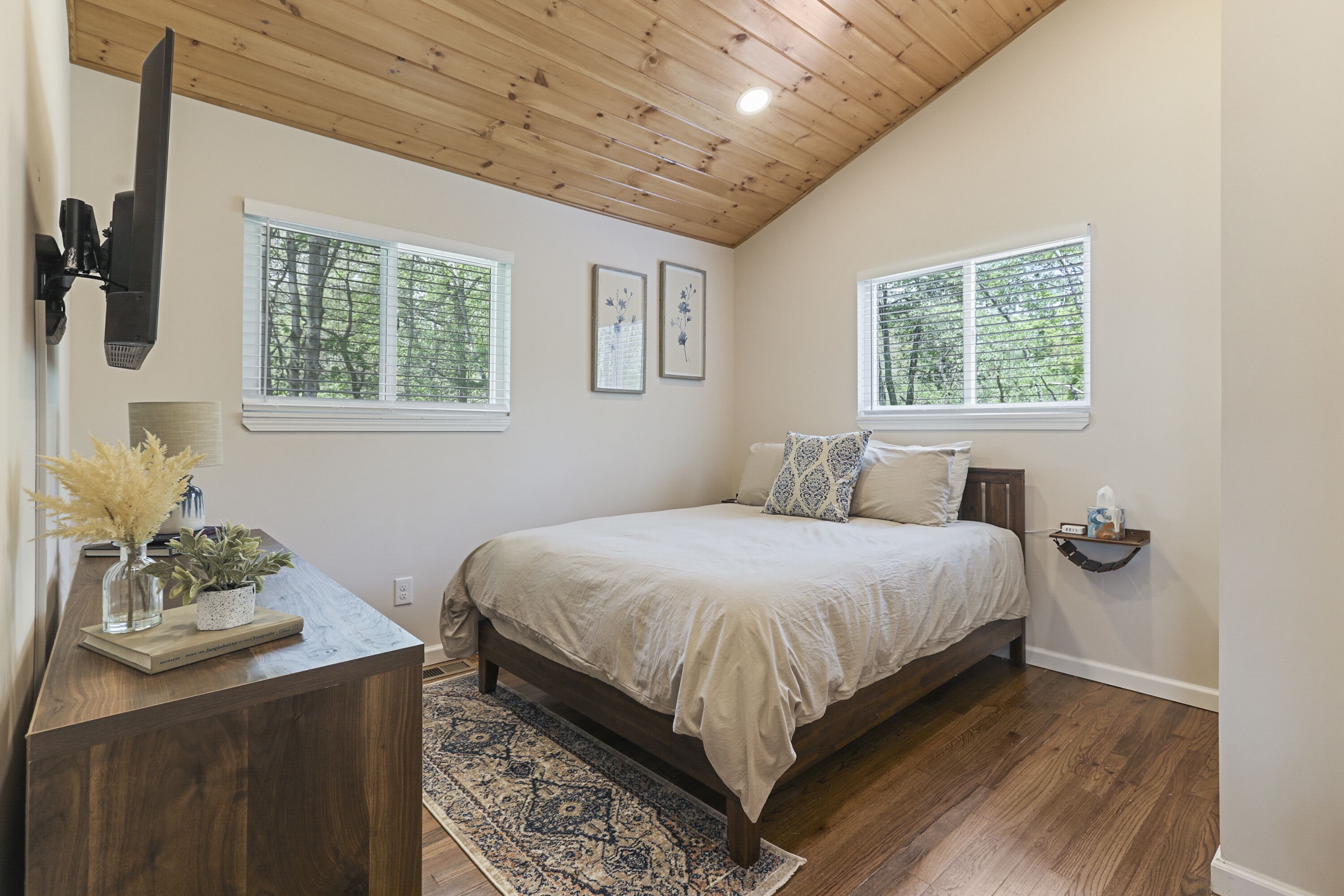A bedroom with a bed, wooden ceiling, two windows with a view of trees, a bedside table with a lamp, and a rug.