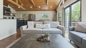 Un vaste espace de vie doté d’un plafond en bois, d’un canapé agrémenté d’un coussin orné de l’inscription « ON PROFITE DU LAC », d’une table basse avec un livre et des télécommandes, et d’une cuisine en arrière-plan.
