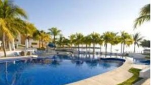 A swimming pool with a curved edge, surrounded by palm trees and a clear sky. - The Hamptons Studio
 (Bucerías)