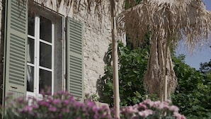 Un edificio rústico con ventana y contraventanas, rodeado de follaje y plantas en flor.