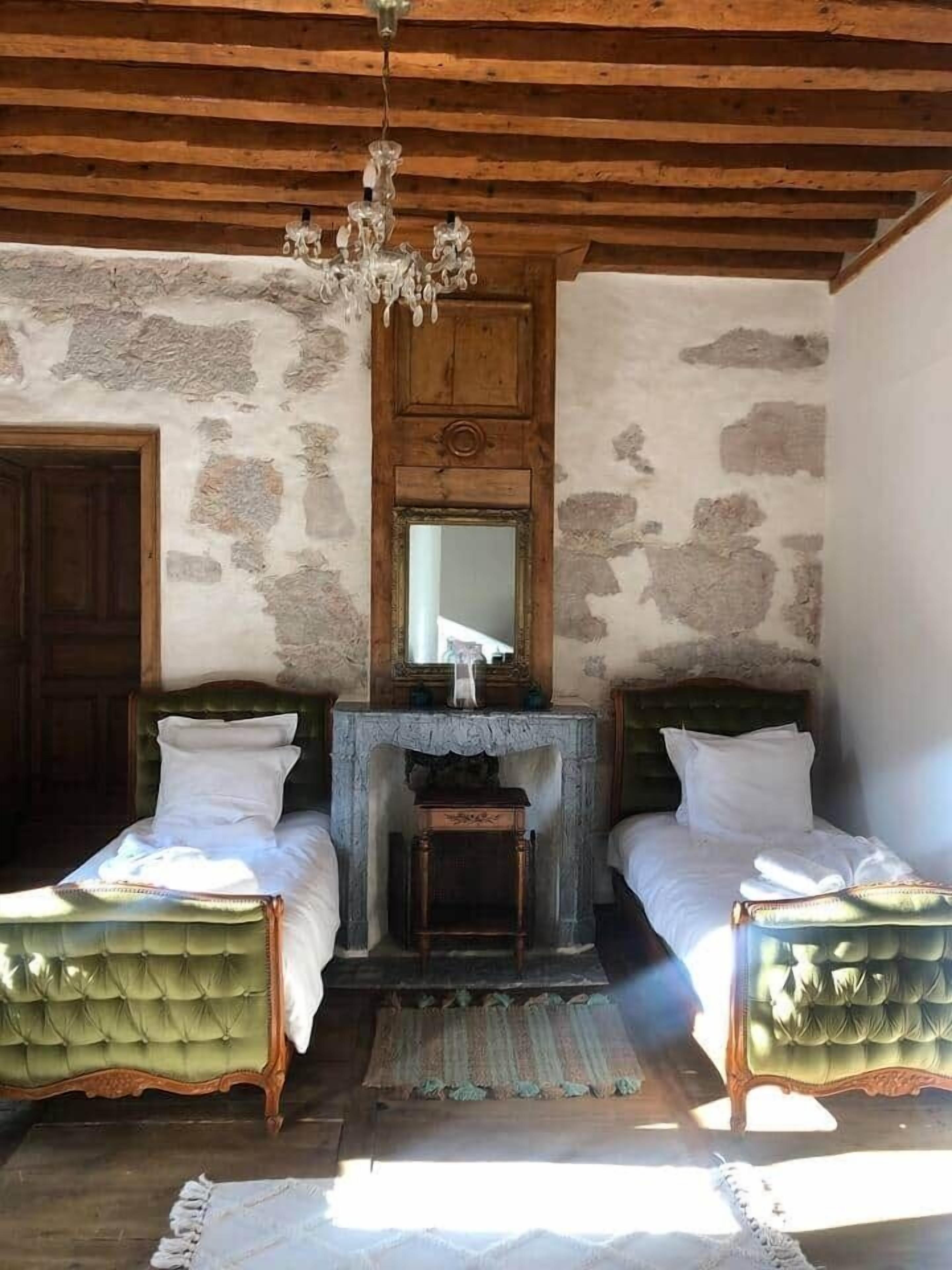 A room with two green upholstered beds, a wooden ceiling, a stone wall, a wooden door, and a mirror above a fireplace.