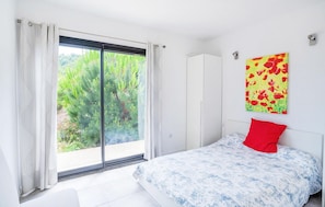 A bedroom with a large sliding glass door leading to a garden, a white wardrobe, and a bed with a blue and white patterned bedspread and a red pillow.
