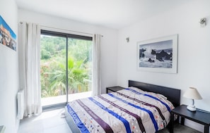 Une chambre avec une grande fenêtre, un lit recouvert d’une couverture à rayures, une table de chevet avec une lampe et un tableau encadré accroché au mur.
