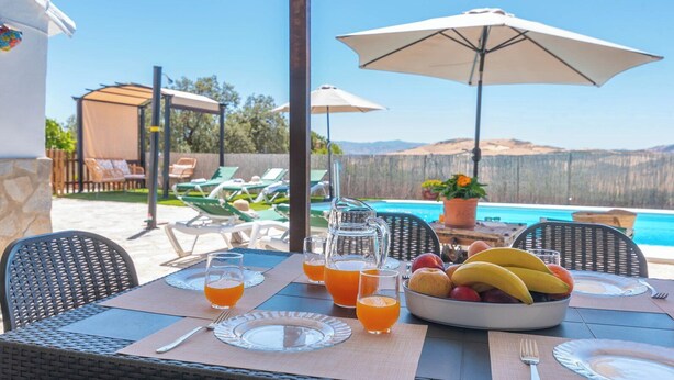 A dining table set with plates, cutlery, and a bowl of fruit, with a pool and lounge chairs in the background. - Casa Pulirio Antequera - La Higuera by Ruralidays (Málaga)