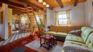 Ein gemütliches Wohnzimmer mit Holzbalkendecke, einer Couch, einem Essbereich mit Schachbrettmuster-Boden und einer Treppe, die in ein weiteres Zimmer führt.