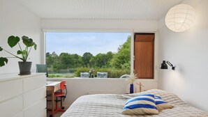 Une chambre à coucher comprenant un lit, un bureau, une chaise, une plante en pot, une fenêtre donnant sur un paysage verdoyant et une lampe.