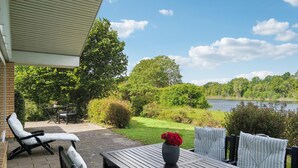Une terrasse extérieure avec une table et des chaises, un vase rempli de fleurs et une vue sur un lac entouré d’arbres.