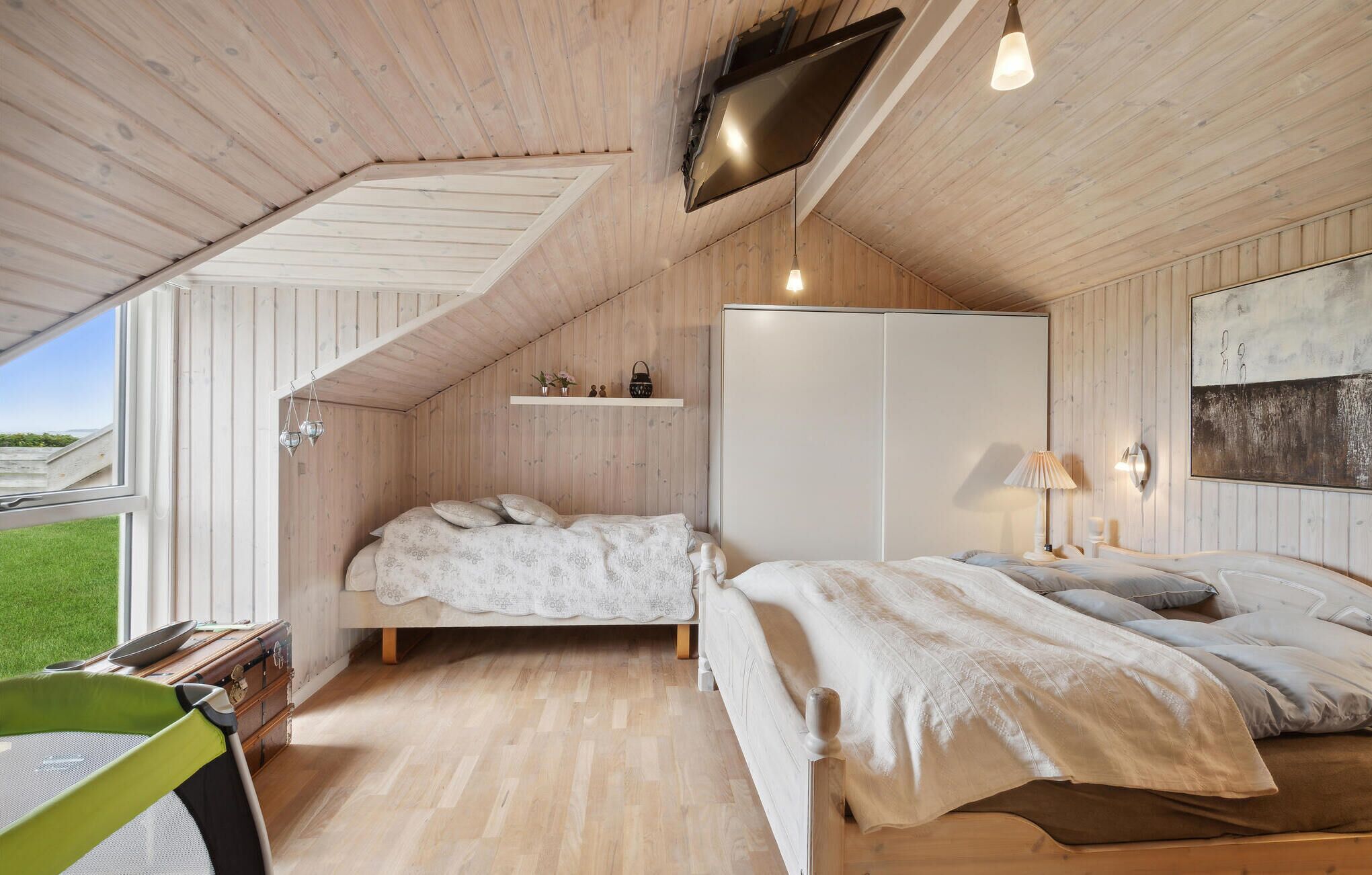 Una camera da letto accogliente con soffitto a falde, un letto, una cassettiera, un televisore montato a parete e una finestra con vista su un paesaggio verde.