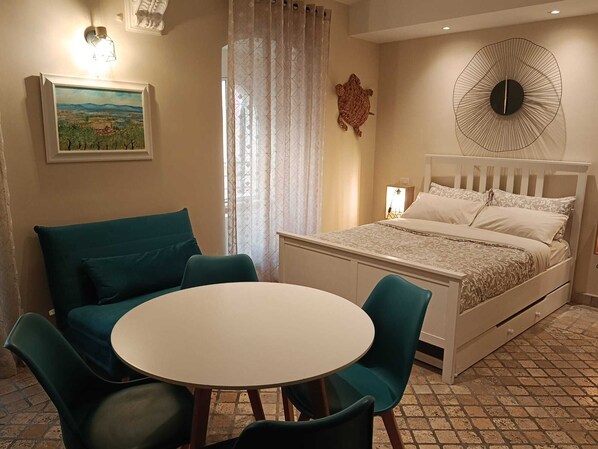 Un dormitorio con una cama, una mesa redonda, dos sillas, un cuadro, un reloj de pared y un adorno de pared.